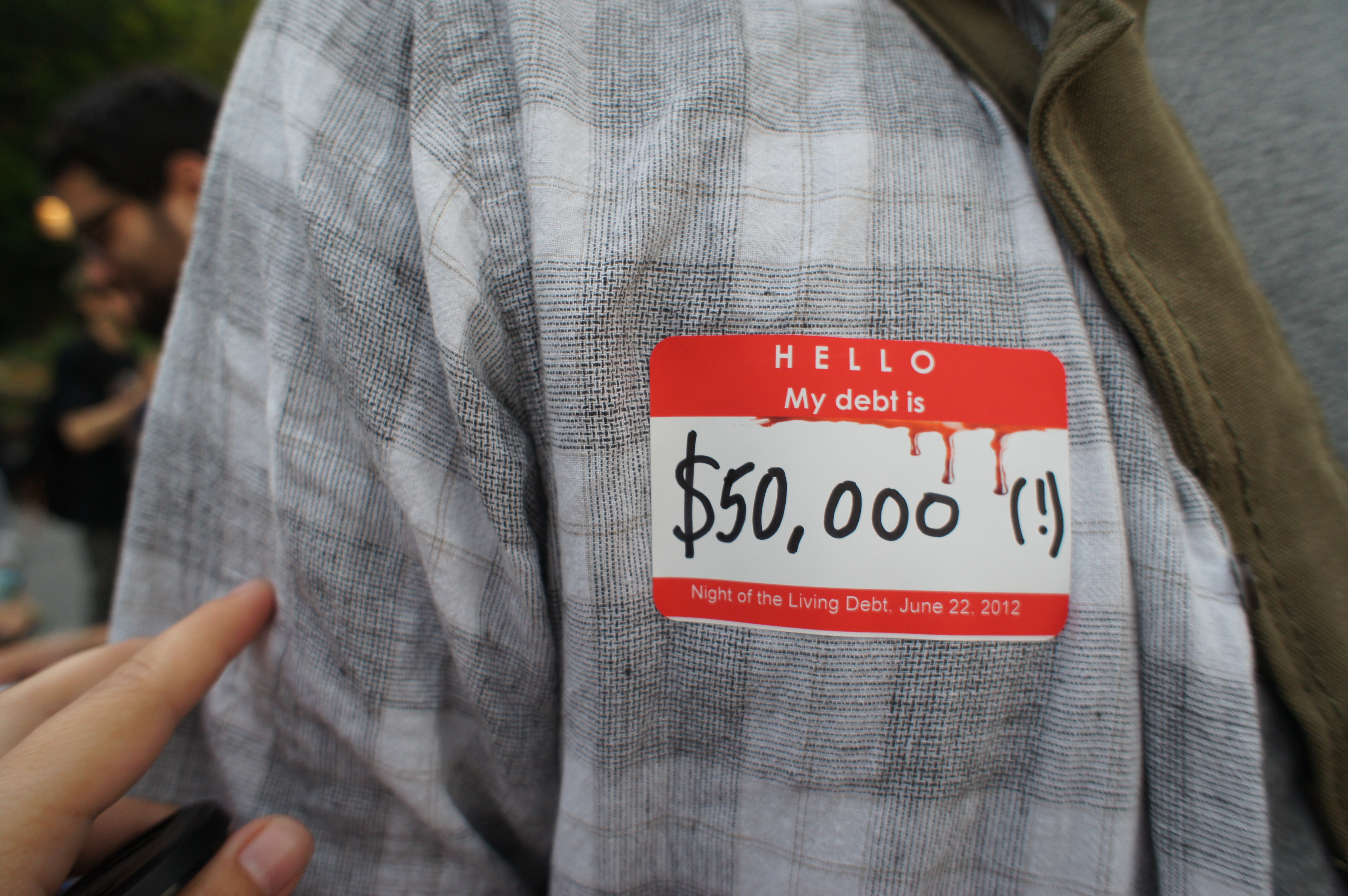 A close-up of a red-and-white name tag on someone’s shirt says, “Hello my debt is $50,000 (!)”