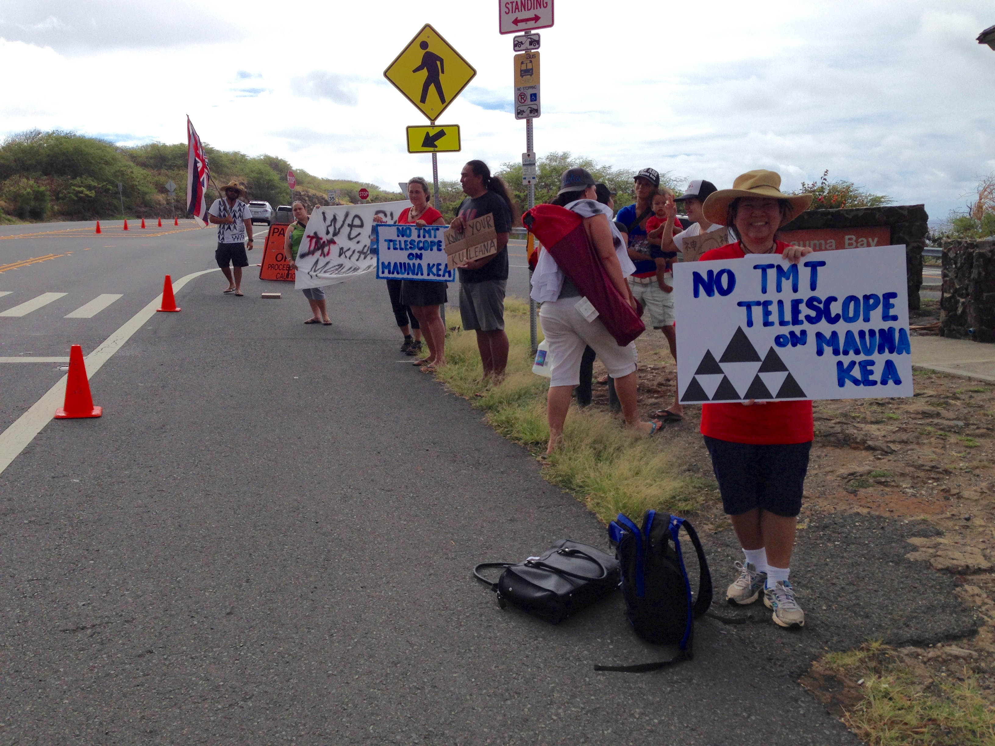 People stand and hold signs on the side of the road. One person in a red shirt holds a sign that reads “NO TMT TELESCOPE ON MAUNA KEA.” The sign is illustrated with triangle-shaped mountains.