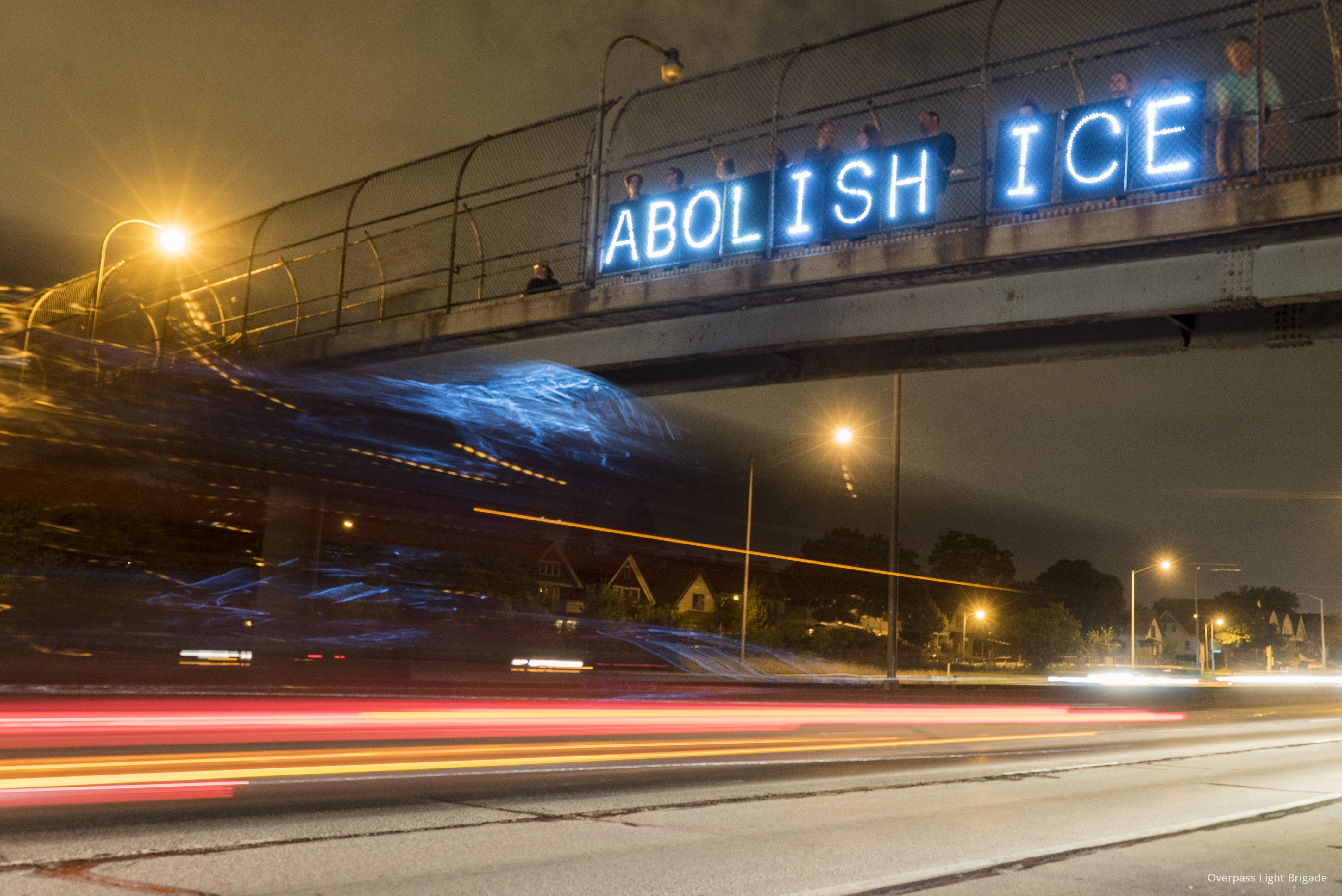 Blurred lights of cars on a highway, photographed at a slow shutter speed. People stand on the overpass above the highway, holding lighted signs that spell out the words “ABOLISH ICE.”