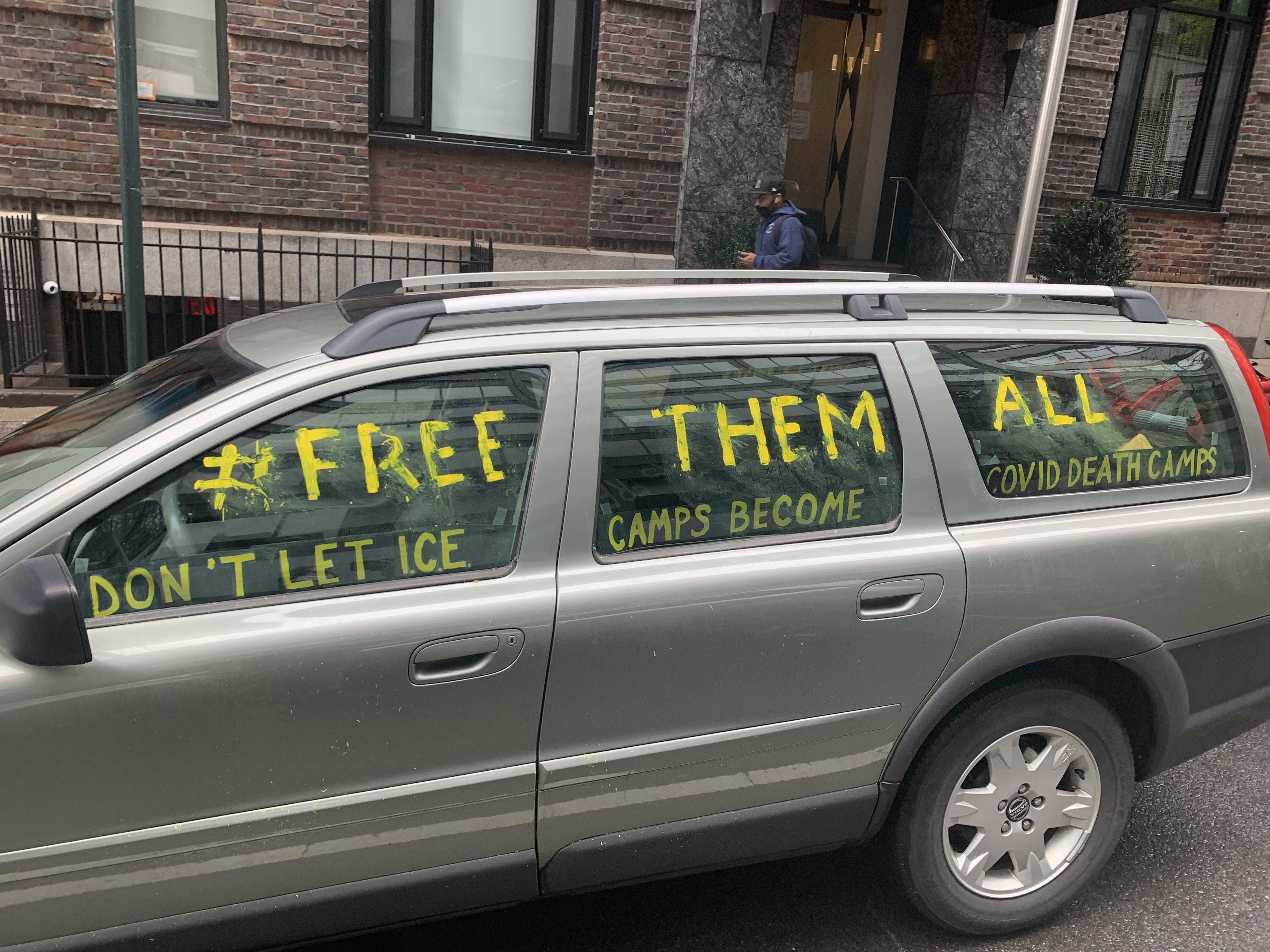 A grey station wagon is parked on the street. Words are painted across the windows in yellow: “#FREE THEM ALL. DON'T LET ICE CAMPS BECOME COVID DEATH CAMPS.”
