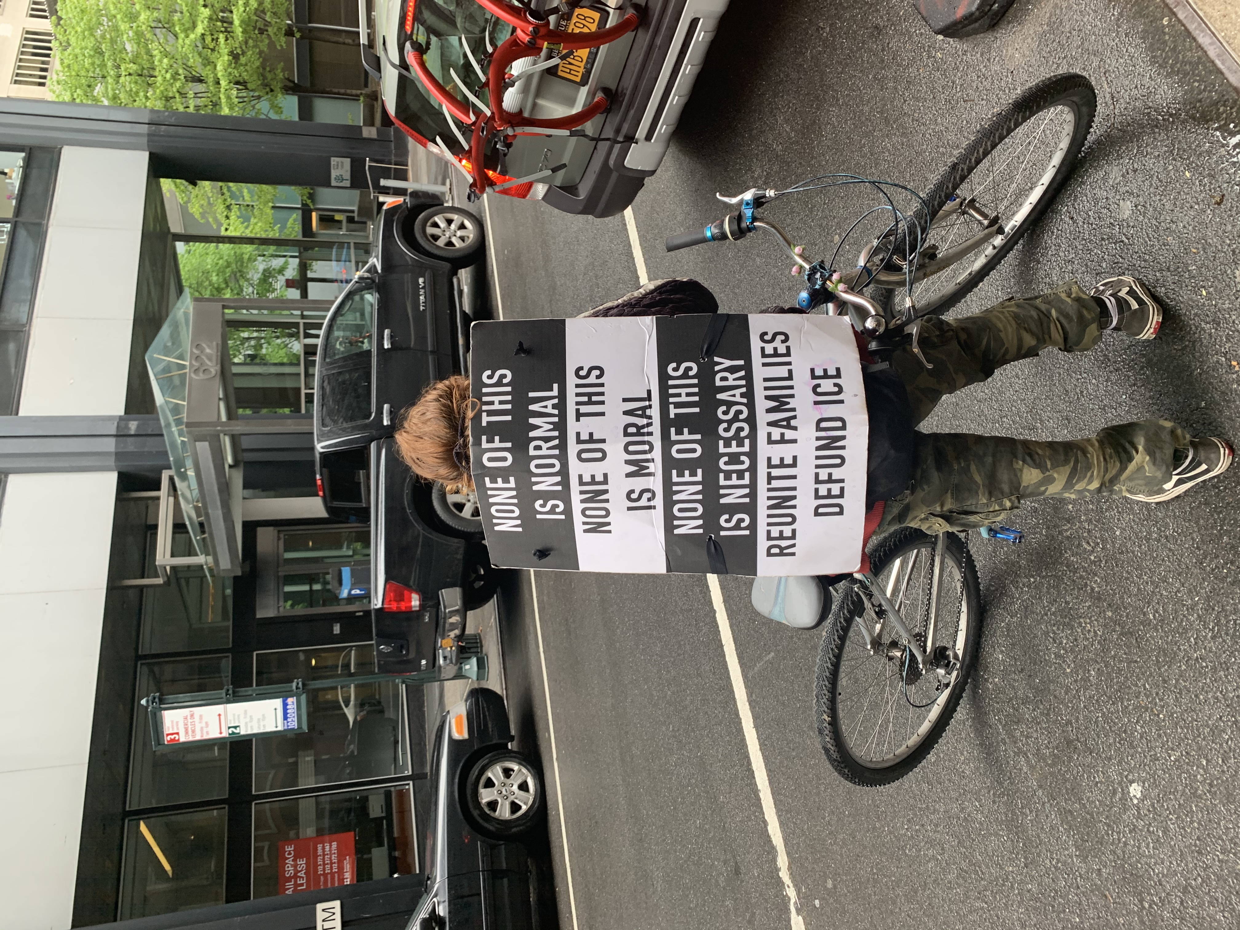 A person stands in a NYC street with a bike. The sign strapped to their back says: “NONE OF THIS IS NORMAL, NONE OF THIS IS MORAL, NONE OF THIS IS NECESSARY, REUNITE FAMILIES DEFUND ICE.”
