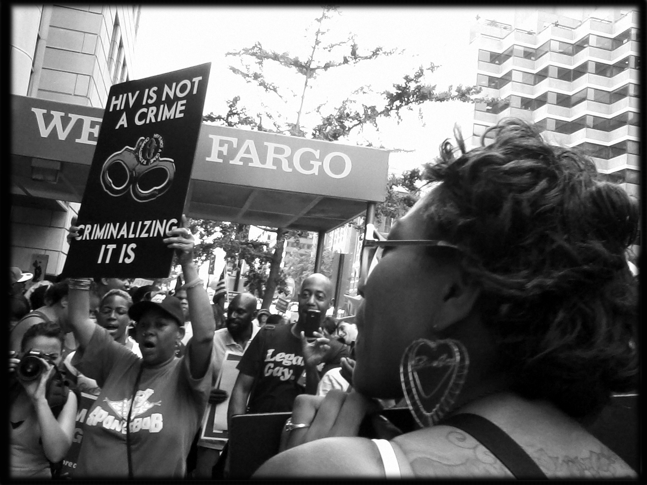 A photograph shows people protesting outside of Wells Fargo bank. One person holds a sign that reads “HIV IS NOT A CRIME - CRIMINALIZING IT IS.” There is a drawing of handcuffs on the sign.