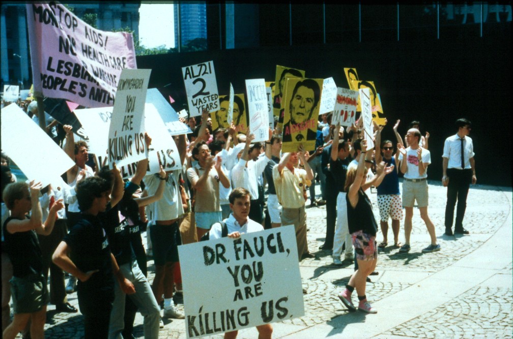 People gather at an ACT UP demonstration in 1990, holding signs. One sign proclaims: “Dr. Fauci, you are killing us.”