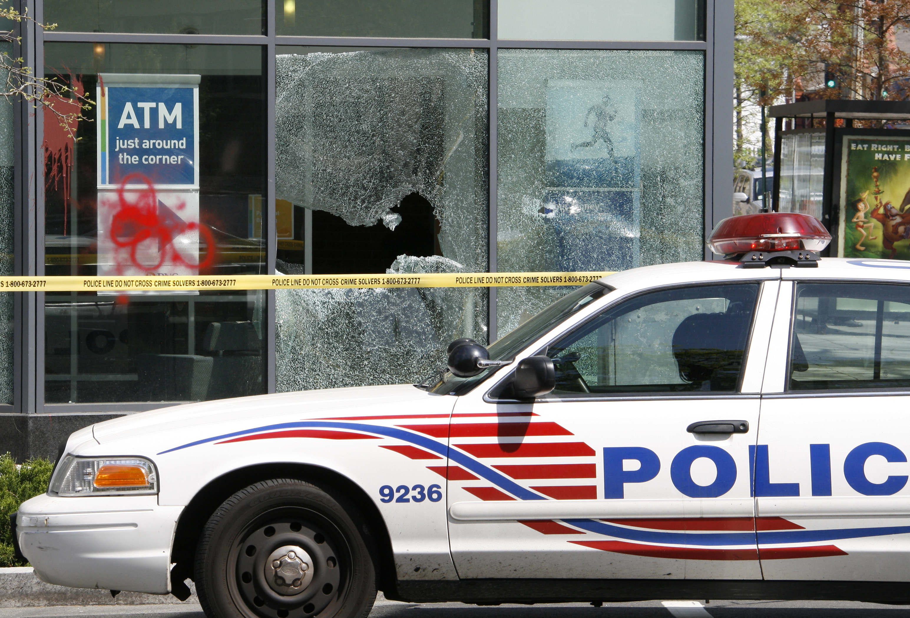 A police car, parked in front of a bank that has had its windows broken and smashed.