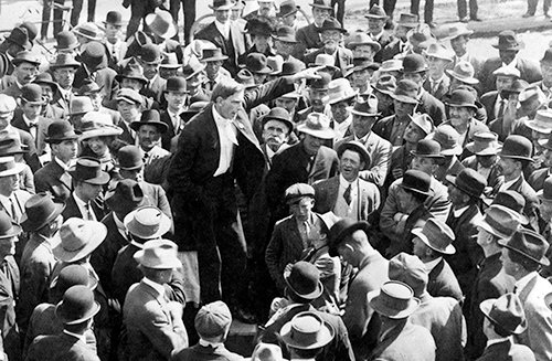 An old black-and-white photograph shows a man, wearing a black suit, who stands on top of a soap box with his arm outstretched and finger pointed. He is surrounded by and addresses a crowd of men who wear bowler hats.