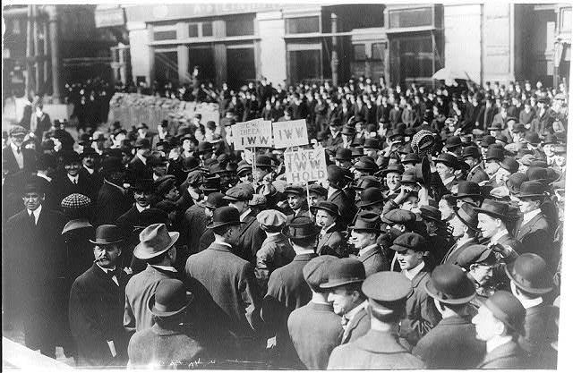 A crowd of people, mostly men, wear bowler hats and collared coats while attending a rally outside a building. A few people are holding signs, which read “TAKE I.W.W. HOLD.”