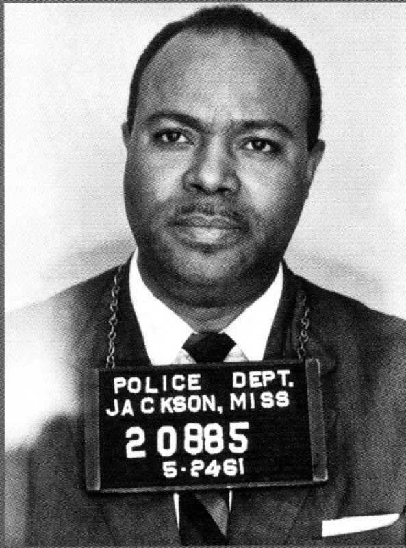 Black-and-white mugshot of Freedom Rider, James Farmer, Jr. Around his neck is a sign, which reads, “POLICE DEPT JACKSON, MISS 20885 5-2461”