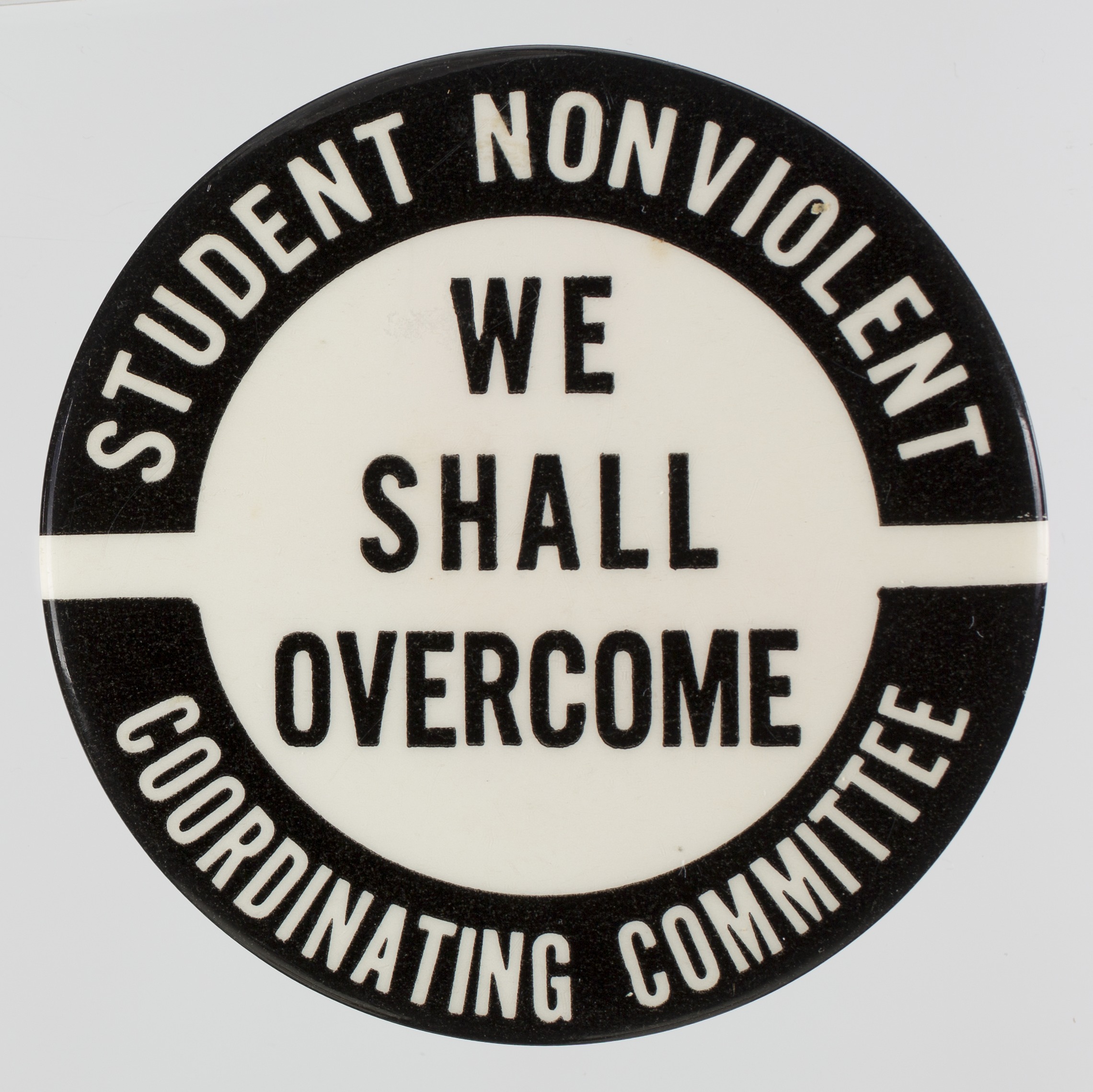 A black and white pin-back button with text. In two arcs around the top and bottom edges of the button face are black blocks with white text. In the middle, centered, is black text. The text reads: [STUDENT NONVIOLENT / WE / SHALL / OVERCOME / COORDINATING / COMMITTEE]. On the reverse are two small white stickers. One is round with the number [225] and the other rectangular with two lines of text: [rear / 6936]. The revers has a pin with a fastener.
