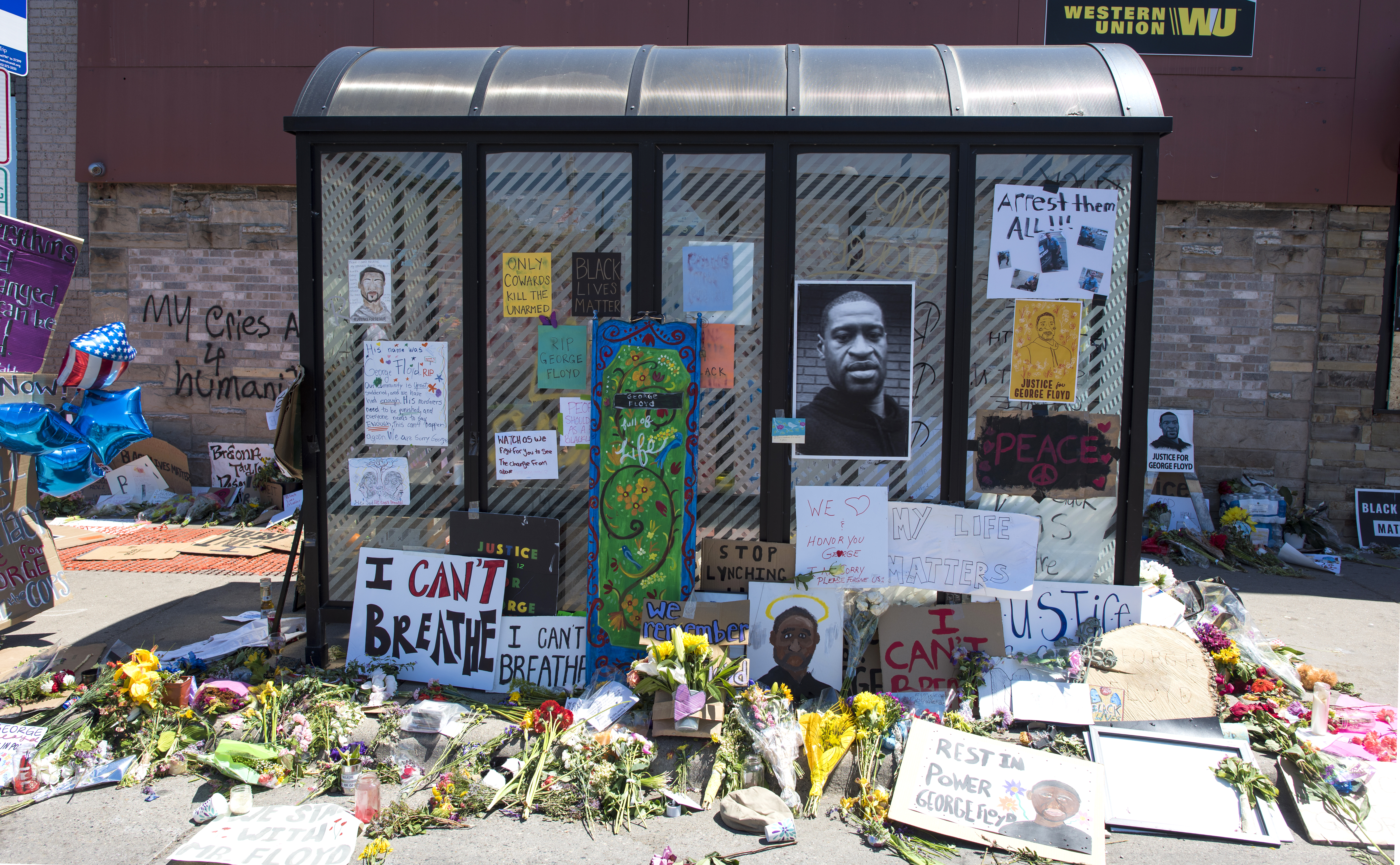 A bus stop shelter, which has been turned into an altar for George Floyd. A photo of Floyd’s face, plus various protest signs, have been taped to the sheter’s windows, and bouquets of flowers cover the sidewalk underneath.