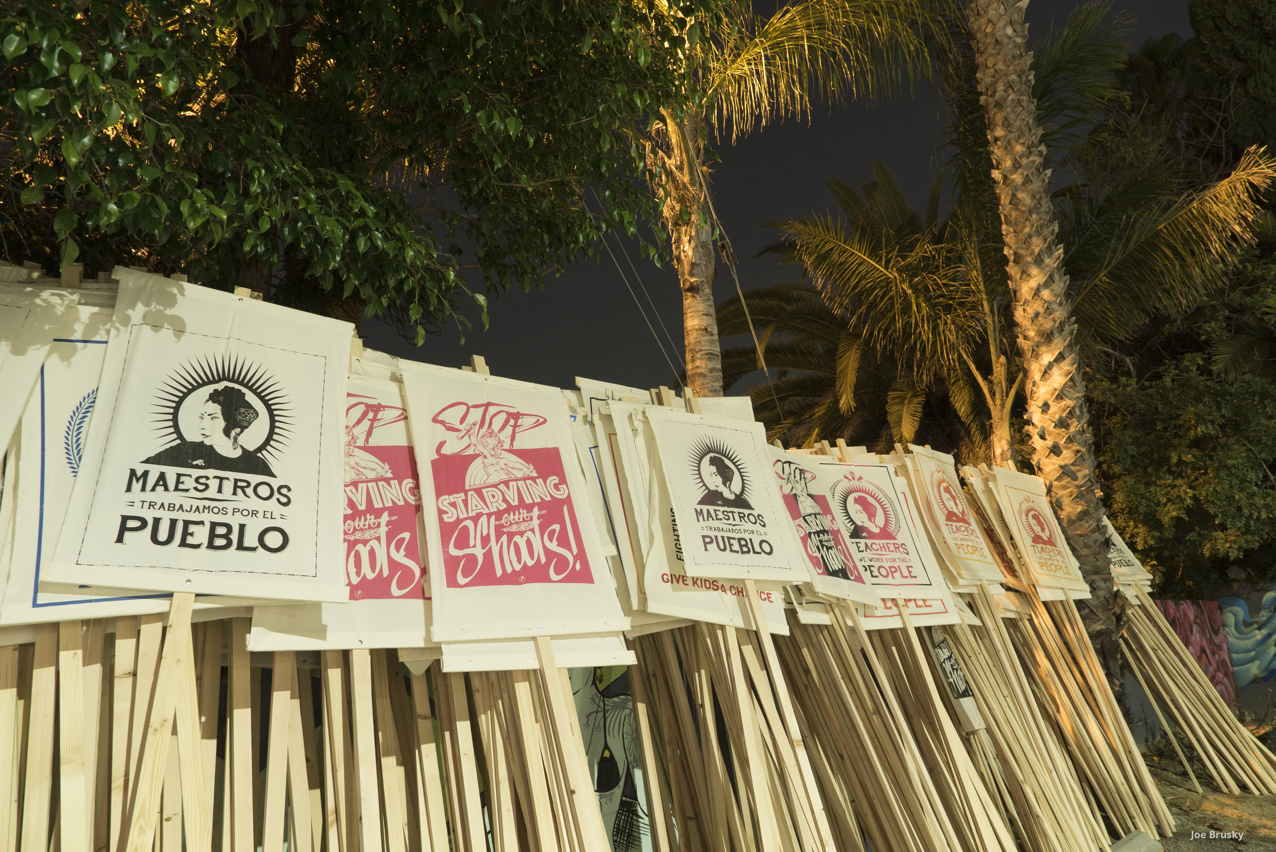 A stack of dozens of picket signs are propped up underneath a canopy of palm trees. The signs are screen printed with the slogans, “Stop starving our schools” and “Maestros Pueblos.” 
