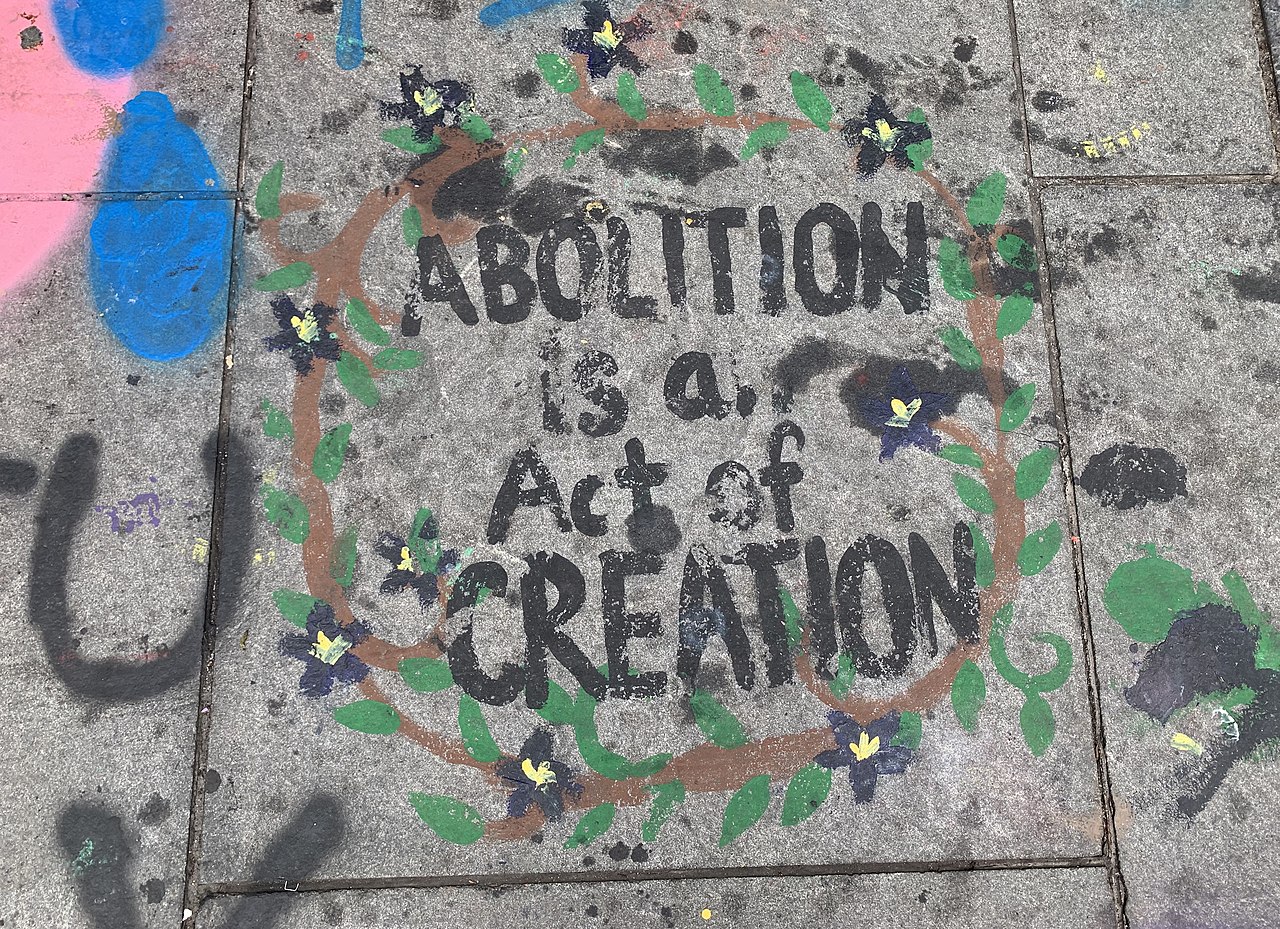 Text in a sidewalk chalk mural reads: "Abolition is a act of creation," and is surrounded in green vines and yellow flowers.